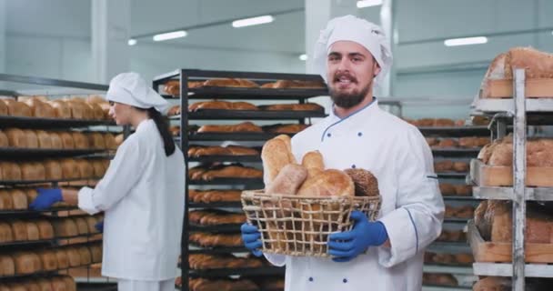 Carismático panadero sonriente con una gran cesta llena de pan recién horneado mirando directamente a la cámara y sonriendo mientras su colega arregla el pan en el estante — Vídeo de stock