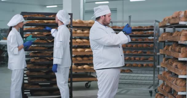 In een bakkerij-industrie hoofd bakker met een nieuwe Tablet neem wat foto's van vers gebakken brood andere twee bakkers op de achtergrond chatten — Stockvideo