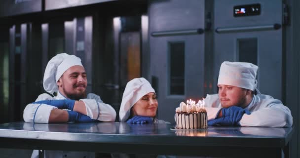 Прекрасная девушка-пекарь в костюме шеф-повара загадывает желание и задувает свечи на торте, после чего ей аплодируют двое веселых мужчин — стоковое видео