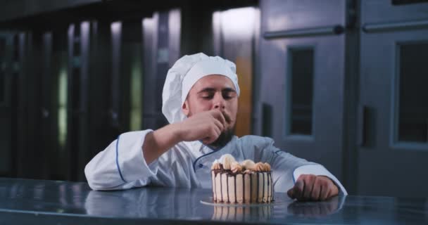 Un hombre guapo y regordete con traje de chefs y barba larga, escoge este pastel de cumpleaños con su dedo y se ve feliz al complacerlo — Vídeos de Stock