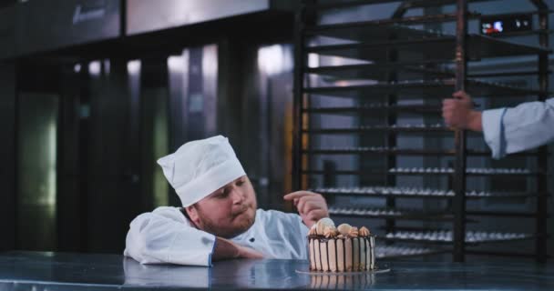 Un joven carismático coge su pastel bellamente decorado con asombro, mientras que un hombre grande empuja una gran bandeja detrás de él — Vídeo de stock