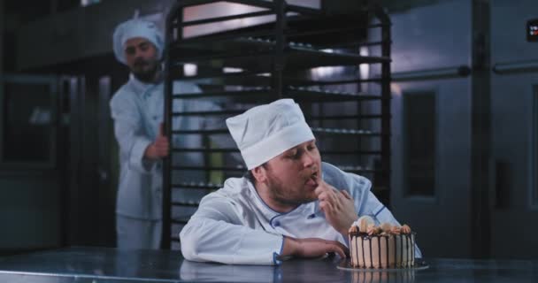 Deux beaux mecs en uniforme de boulanger regardent ce gâteau riche et appétissant et le goûtent avec étonnement au goût étonnant, ils en discutent tous les deux — Video