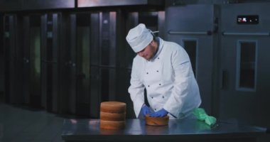 Genç bir tombul görünümlü şef yarısında bir pasta açık keser, o yanında yığılmış diğer üç kek ve arka planda bir fırıncı çalışma alanı varken