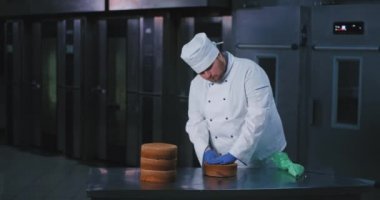 Uygun şefler kıyafetleri ile bir husky iyi görünümlü fırıncı adam yarısında pasta keser ve arka planda fırınlar olduğu gibi tutarlılık için kontrol