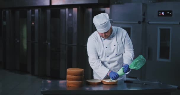 Un hombre barbudo grande con un uniforme de panaderos, guantes azules y sombrero de chefs, decoró el interior de la torta con hielo blanco esponjoso que viene de la bolsa de tuberías, mientras que hay un montón de hornos grandes en el — Vídeo de stock