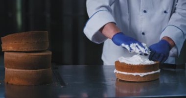 Mavi lastik eldiven giyen tombul elleri olan bir fırıncı eliyle pastanın üzerine ipeksi beyaz krem sarıyor.
