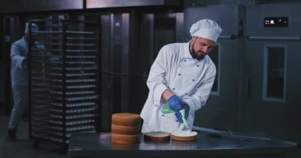 Un hombre atractivo con barba con un traje de cocina termina de decorar el interior del pastel, mientras que un hombre gordo con barba de barba camina detrás de él rodando una bandeja — Vídeo de stock