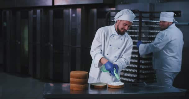 Un joven chef atractivo y regordete decoró el interior de la torta con crema blanca y esponjosa proveniente de una bolsa de tuberías verde claro, mientras que otro panadero barbudo grande se detuvo y tuvo una pequeña charla con — Vídeos de Stock