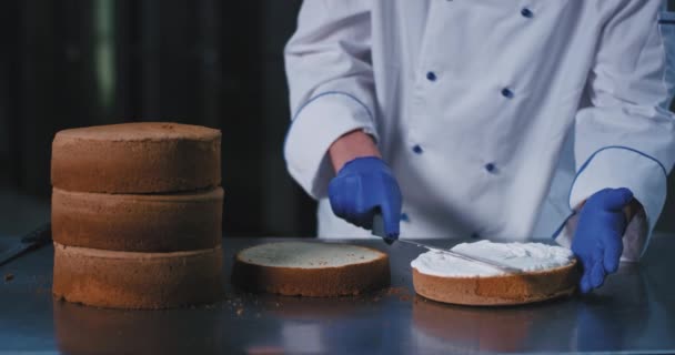 Een close-up van een paar blauwe handschoenen vasthouden van een spatel en sierlijk verspreiden van de crème over de cake, zodat het ziet er glad — Stockvideo