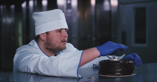 Um gordinho enfrentou encantador padeiro pegou a cereja vermelha de um bolo e está olhando para ele hipnotizado — Vídeo de Stock