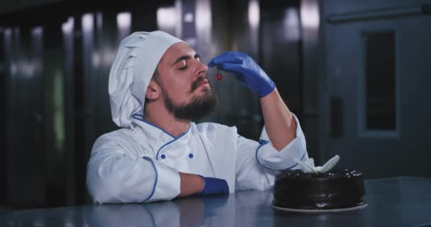 Un joven panadero fuerte y guapo miró curiosamente la cereza roja antes de colocarla cuidadosamente en el pastel de chocolate y mirar a la cámara con felicidad mientras sonreía. . — Vídeo de stock