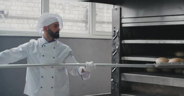 Working hard good looking baker with a chin and stylish uniform take off the cooked bread from oven using a torch his assistance take the bread and arrange them on the shelves — Stock Video