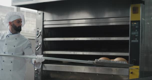 Panadería profesional de la fábrica de panadería chef en un uniforme especial chef sacar el pan de la máquina del horno su ayuda a organizar en los estantes — Vídeos de Stock