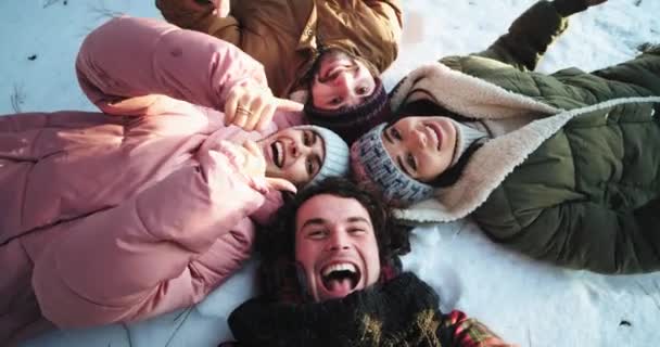 Grupo animado de amigos deitados no chão nevado no meio da floresta e aproveitando o tempo juntos fazem um vídeo engraçado usando uma câmera para selfies — Vídeo de Stock