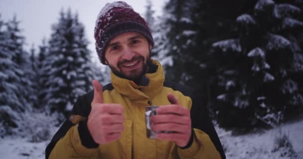 Гарно виглядає турист посеред лісу в зимовий день, дивлячись прямо на камеру, п'ючи гарячі напої, які посміхаються великими і відчувають захоплення. повільний — стокове відео