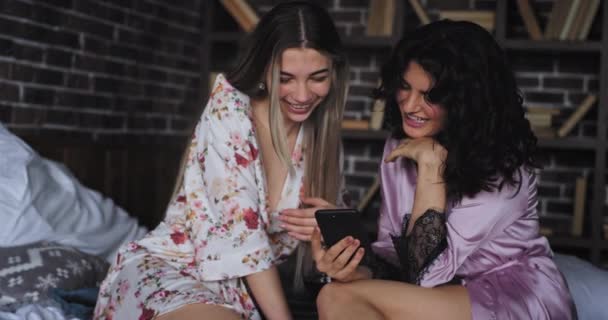 Улыбаясь, две харизматичные женщины в пижаме проводят время вместе, наблюдая через смартфон взволнованный и счастливый, сидя на кровати — стоковое видео
