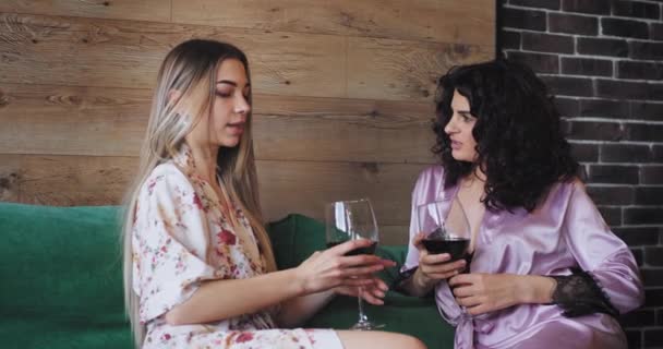Multi etniczne panie bardzo charyzmatyczny ciesząc się wieczorem razem z kieliszków wina w piżamie na kanapie rozmawiają ze sobą — Wideo stockowe