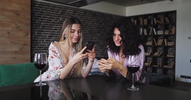 多民族的两个朋友女士穿着睡衣在厨房酒吧桌子上,用酒杯看着通过智能手机微笑大,互相聊天 — 图库视频影像