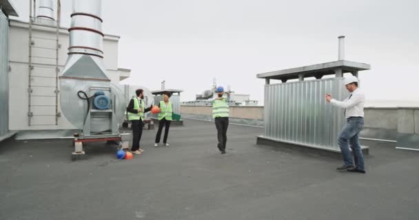 Ein Mann mit blauem Bauhut und neonfarbener Jacke unterhält die anderen Arbeiter mit seinen Bewegungen, während sich hinten ein gutaussehender Mann und eine gut aussehende Frau Getränke teilen. — Stockvideo