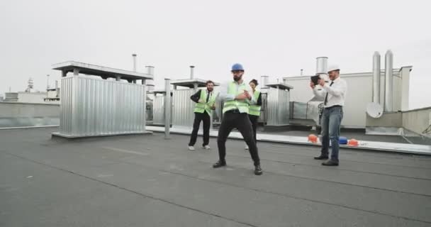 Трое строителей, красивый мужчина и красивая женщина медленно танцуют за парнем, который танцует профессионально, пока снимают на архитектурной площадке — стоковое видео