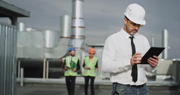 The young construction man presses on his iPad with his index finger while two good looking architect people pass him by in deep conversation — Stock Video