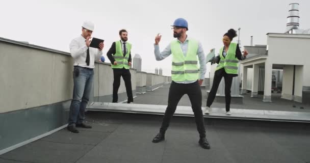 Drei Bauarbeiter tanzen zusammen und genießen sich gegenseitig, sie stehen auf einer Baustelle und tanzen mit, während ein gut aussehender Mann in einem atemberaubenden weißen Hemd und Krawatte sie filmt. — Stockvideo