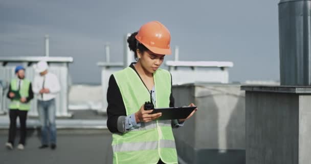 A jovem senhora da construção de pele mista parece muito focada em seu trabalho como ela parece surpreso com seu iPad, ela é muito linda, e parece bonito vestindo um capacete de construção laranja — Vídeo de Stock