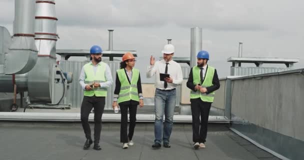Zwei Geschäftsleute und ein junger Ingenieur mit Architekt analysieren den Plan der Baustelle und gehen mit einem Tablet durch das Dach des Gebäudes — Stockvideo