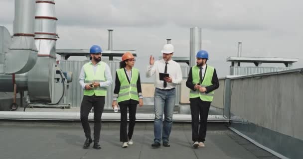 Dos hombres de negocios y un joven ingeniero con arquitecto analizando el plan de construcción y utilizando una tableta que caminan por la azotea del edificio — Vídeos de Stock