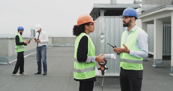 Két mérnökök a tetőn építkezési hely többnemzetiségű emberek egy beszélgetést a terv az épület háttér két üzletember vesz néhány képet, és megjegyzi, — Stock videók