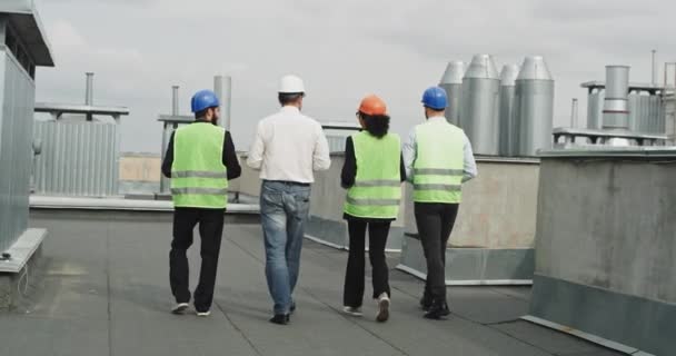 Em um edifício moderno telhado Africano senhora engenheiro assistência e arquitetos analisando o plano de construção andando por aí usando capacetes de segurança — Vídeo de Stock