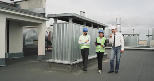 Dos arquitectos hombres y su ayuda mujer africana analizando el plan de construcción caminando en la azotea de la construcción sosteniendo una tableta tienen una discusión concentrada — Vídeos de Stock