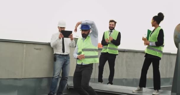 Charismatische ingenieur met een baard dansen op de pauzetijd voor zijn collega's op het dak van de bouwplaats ze genieten van de tijd perfect team — Stockvideo