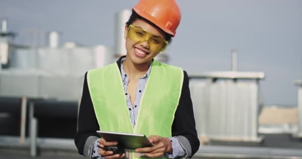 Красивая африканская этническая молодая женщина-архитектор смотрит прямо в камеру и улыбается, держа в руках рацион и планшет, и иметь оборудование безопасности — стоковое видео