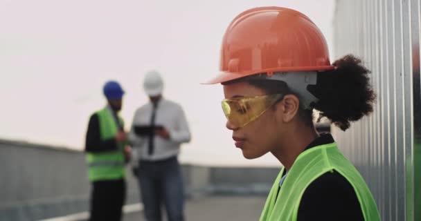 Charismatische jonge vrouw ingenieur Afrikaanse etnische drinken wat water op het dak van de bouwplaats ze het dragen van een veiligheidsuitrusting oranje helm en gele bril — Stockvideo