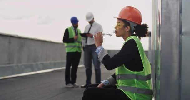 在建筑工地上，美丽的微笑的非洲女工程师在建筑屋顶上喝着瓶子里的水，她戴着安全帽和黄色安全眼镜 — 图库视频影像