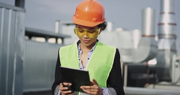 Impresionante mujer joven ingeniero analizando el plan de la obra desde la tableta hablando de la ración con otro trabajador y el uso de casco de equipo de seguridad y gafas amarillas — Vídeos de Stock