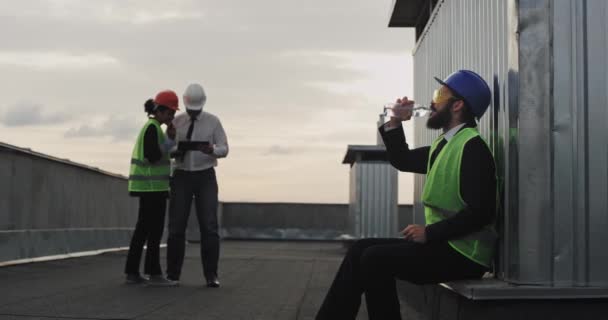 Man ingenieur met een baard en veiligheidsuitrusting op bouwplaats drinkwater uit een plastic fles glimlachend schattig naar de camera zijn collega's werken geconcentreerd op de achtergrond op — Stockvideo