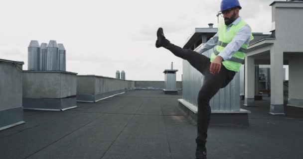 Харизматичный мужчина в шлеме на стройке танцует молодой инженер с бородой, взволнованный на крыше здания — стоковое видео