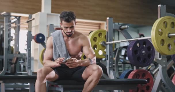 En tilltalande ung man dricker vatten medan du tränar, han ser väldigt trött ut som han försöker att upprätthålla ett hälsosamt liv — Stockvideo