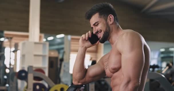 Joven con un cuerpo en forma hablando por teléfono en una gran clase de gimnasia después de sus ejercicios de entrenamiento tomar un descanso — Vídeos de Stock