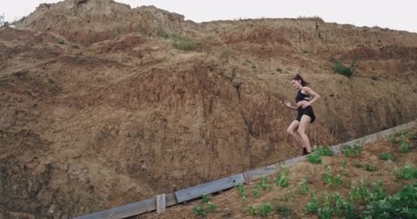 Una joven de buen aspecto corre por un tramo de escaleras en la naturaleza y se acerca a una playa que está haciendo deporte y ser saludable . — Vídeo de stock