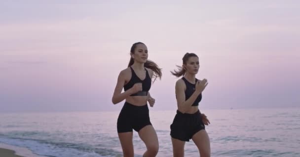 美丽的两位女士在海滩边奔跑，集中享受着早晨练习运动的时刻。慢动作 — 图库视频影像