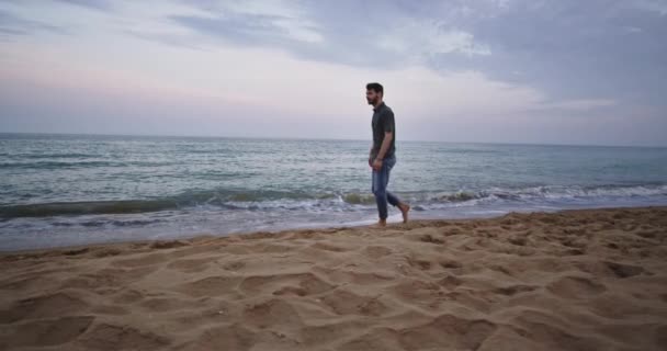 Um homem bonito com uma barba caminha na praia, onde as ondas estão e ele olha misteriosamente ao redor — Vídeo de Stock