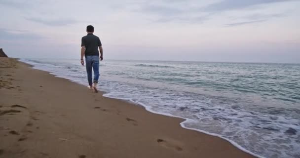 Een fit jonge man loopt op het strand met zijn blote voeten, hij ziet er erg geïnintrigeerd door de golven en hij kijkt naar het landschap — Stockvideo