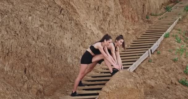 Dwie panie w czarnej odzieży sportowej rozciągające nogi na drewnianych stentów obok tła plaży duże skały — Wideo stockowe