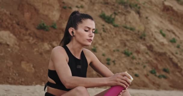 Fáradt hölgy iszik szomjas vizet a strandon ülve, utána kemény edzés reggel a tenger mellett — Stock videók