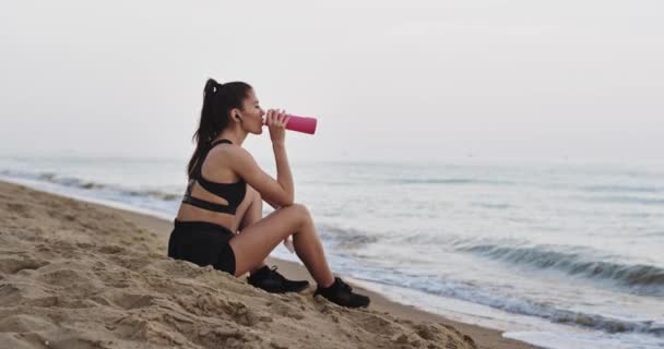 Am Morgen nach dem Training eine Pause am Strand vor dem Meer einlegen, Wasser trinken und die Stille genießen — Stockvideo