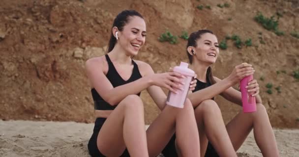 Sonriendo grandes damas atractivas en una ropa deportiva en la playa frente al mar bebiendo agua sedienta de una botella de plástico — Vídeos de Stock