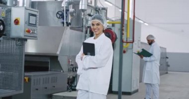 Özel beyaz üniforma giyen bir elektronik tablet tutarken büyük bir imalat sektöründe Heyecanlı olgun kadın mühendis kameraya doğrudan bakıyor